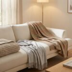 Un salon moderne et lumineux avec un canapé beige décoré de trois plaids texturés. Une table basse en bois, un livre et une plante.