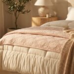 Lit aux draps doux et couettes superposées dans les tons beiges et roses, avec une couverture tricotée. Lumière chaleureuse.