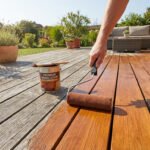 Une main applique de la teinture bois sur une terrasse, transformant le bois gris vieilli en une surface riche et brillante.