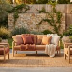 Salon de jardin organique avec canapé et deux fauteuils en bois clair, coussins et plaids aux tons terre. Tapis en jute, plantes en pot.