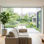 Salon moderne avec canapé beige, grande plante et larges baies vitrées s'ouvrant sur un jardin luxuriant et paysagé.