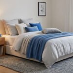 Lit confortable avec des coussins bleus et neutres, table de chevet en bois et tapis gris dans une chambre lumineuse.