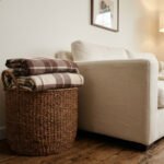 Un panier en osier rempli de plaids à carreaux marron placé à côté d'un canapé beige dans un salon chaleureux.