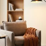 Un fauteuil gris confortable avec un plaid, une lampe sur pied et une étagère remplie de livres près d'une fenêtre.