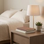 Table de chevet en bois avec lampe, livres empilés, petite plante succulente et un verre d'eau, près d'un lit douillet.