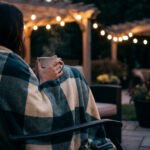 Femme enroulée dans un plaid à carreaux, tenant une tasse chaude sur une terrasse éclairée par des guirlandes.