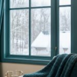 Une personne portant des gants fermant un rideau épais devant une fenêtre givrée avec un paysage enneigé en arrière-plan.